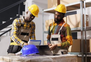 Logistics managers analyzing inventory and comparing data on laptop and clipboard in warehouse. African american men checking goods supply schedule and working in distribution department storehouse