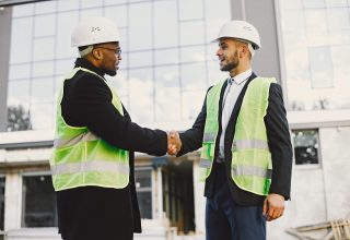 Multi racial builders handshaking outdoors. Wearing uniform, talking about new glass building. Working on the poject. City infrastructure. Black man.